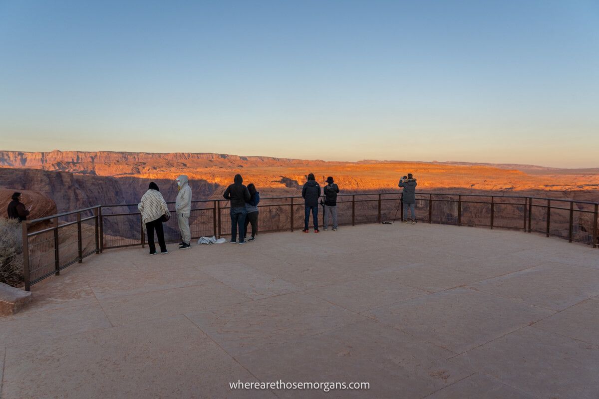 Large paved viewing area fenced in for protection from falling over the edge of cliffs with the sun rising over a flat rocky plateau ahead