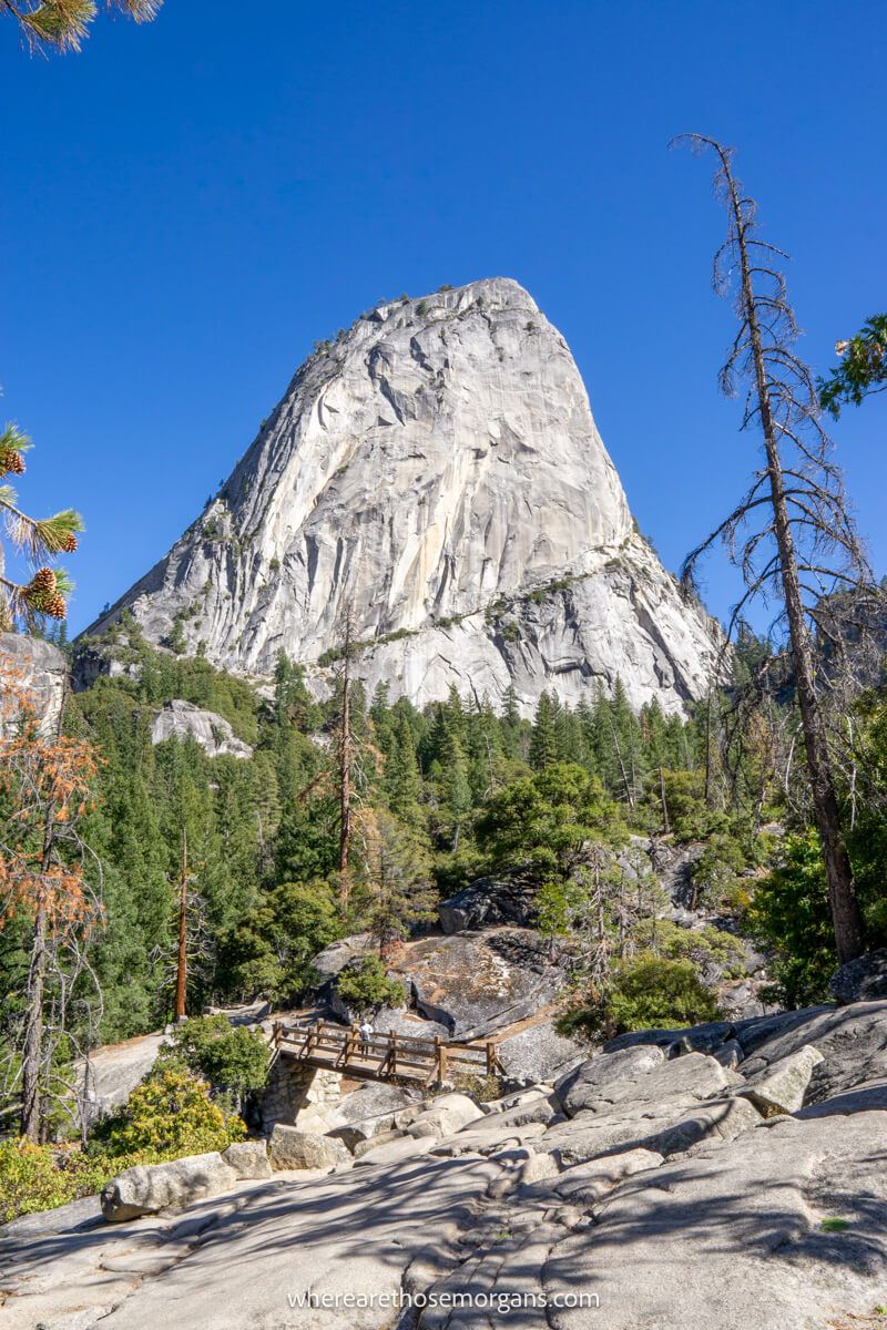 Rocky hiking trail leading to a wooden bridge surrounded by evergreen trees and a tall granite monolith in the distance on a clear day with blue sky on the Mist Trail in Yosemite