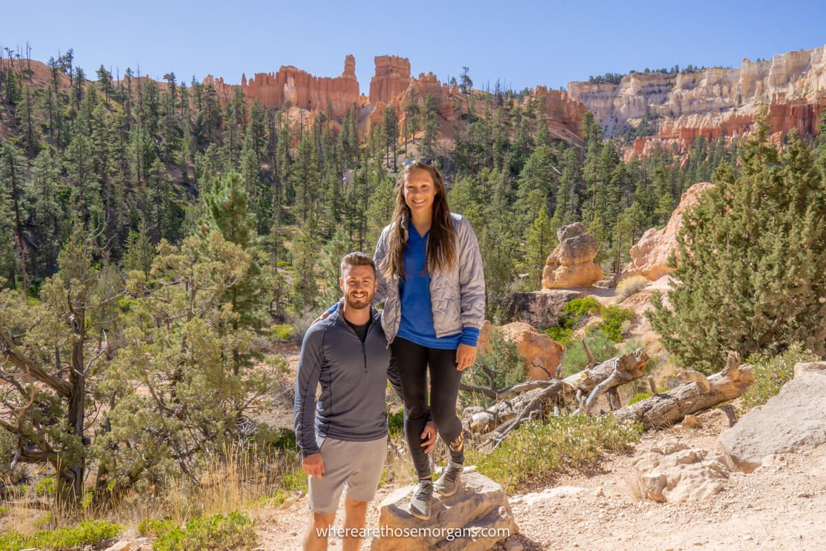 Two hikers stood together for a photo with views behind of trees and sandstone cliffs on a bright day