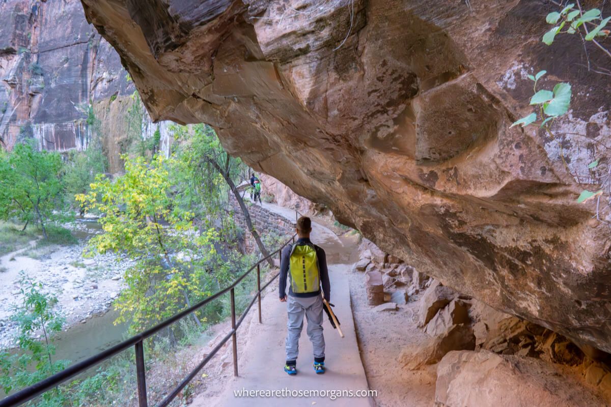 Hiker with waterproof backpack and pants walking along a narrow trail next to a fence and overhanging red rock cliff