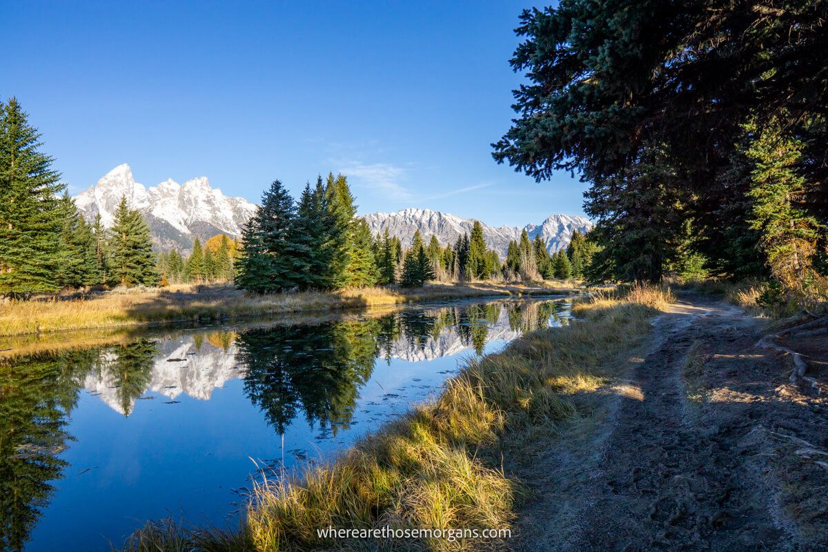 Hiking path in shadow alongside a still river with trees and mountains reflecting at Schwabacher Landing in Grand Teton National Park