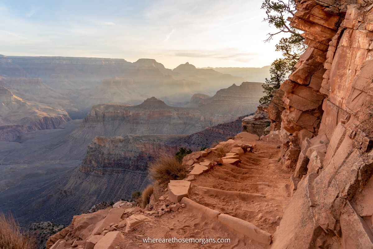 Photo of the dusty, rocky and narrow South Kaibab hiking trail next to a tall cliff wall leading down into the vast Grand Canyon with intense beams of light bursting into the canyon at sunrise