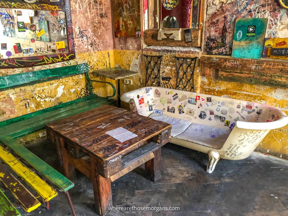 Room in a ruined building with a bathtub next to a table and bench, writing on the walls and a mirror