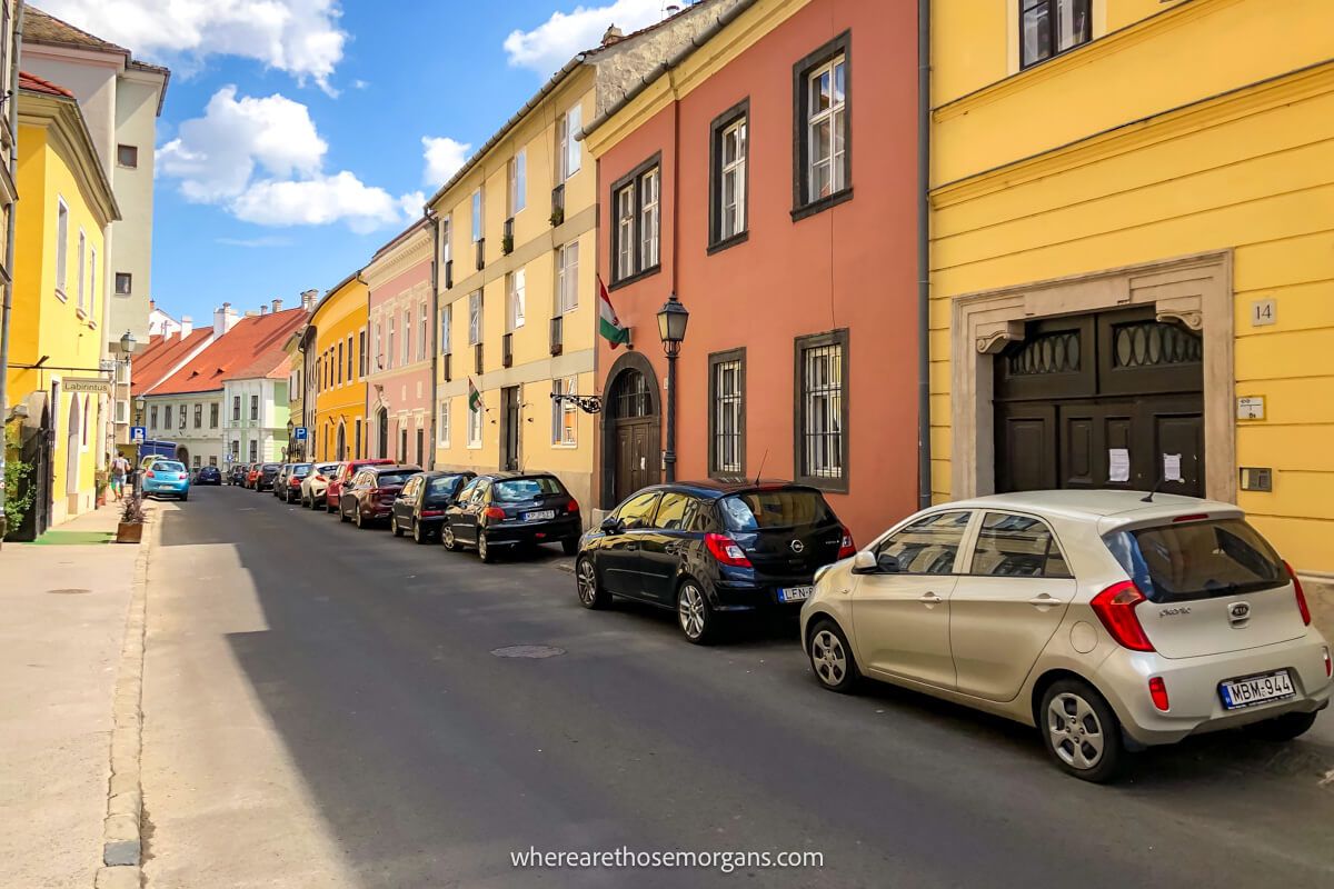 Photo of a narrow street with a long line of houses joined together painted yellow and orange with cars parked outside on the street