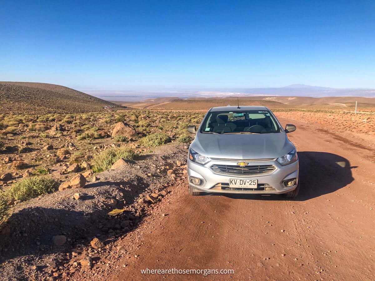 Small silver car parked on a dirt road in a wide open landscape with green vegetation and big blue sky