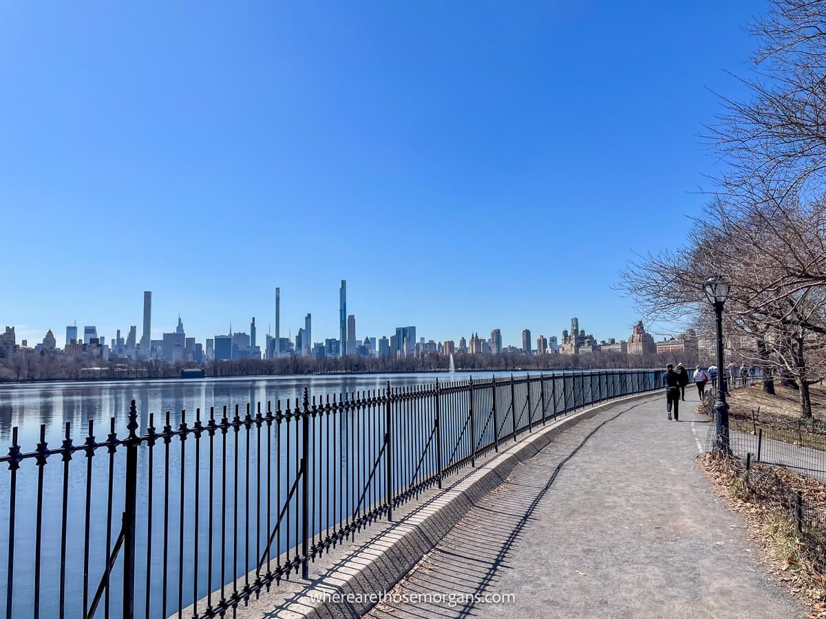 Walking path and huge reservoir fenced off by a tall black gate with city views in the distance