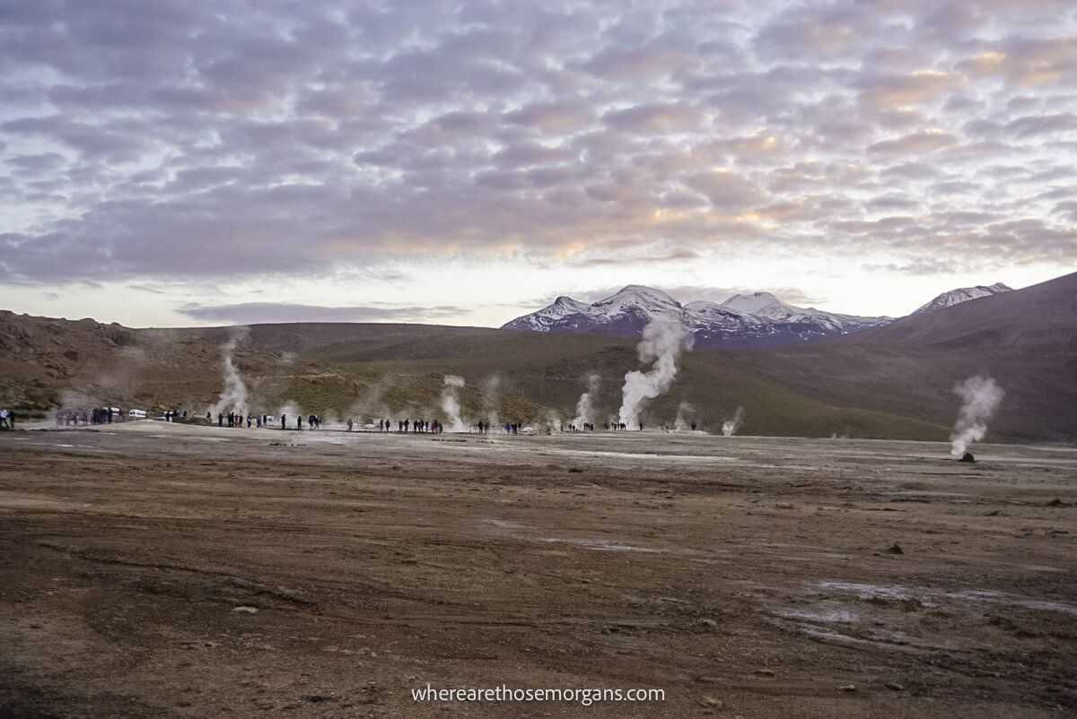 Photo of a wide open field with lots of lightly billowing geysers in front of tall mountains with clouds in the sky