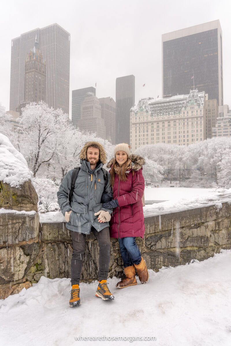Photo of a couple standing together in heavy winter coats with hoods up on a low stone bridge with views of NYC behind in the snow