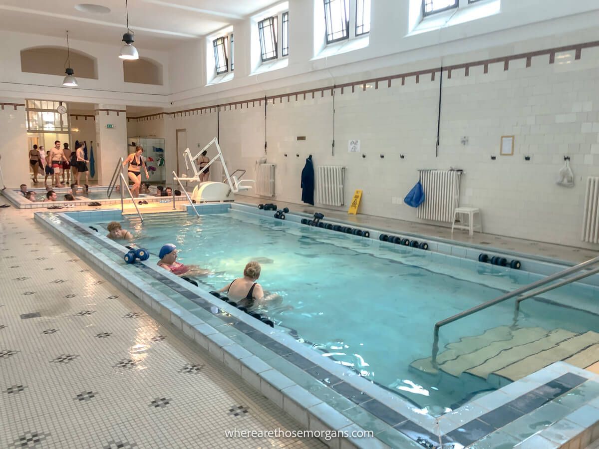 Photo of an indoor pool with bathers sat talking and others walking into a white tiled room