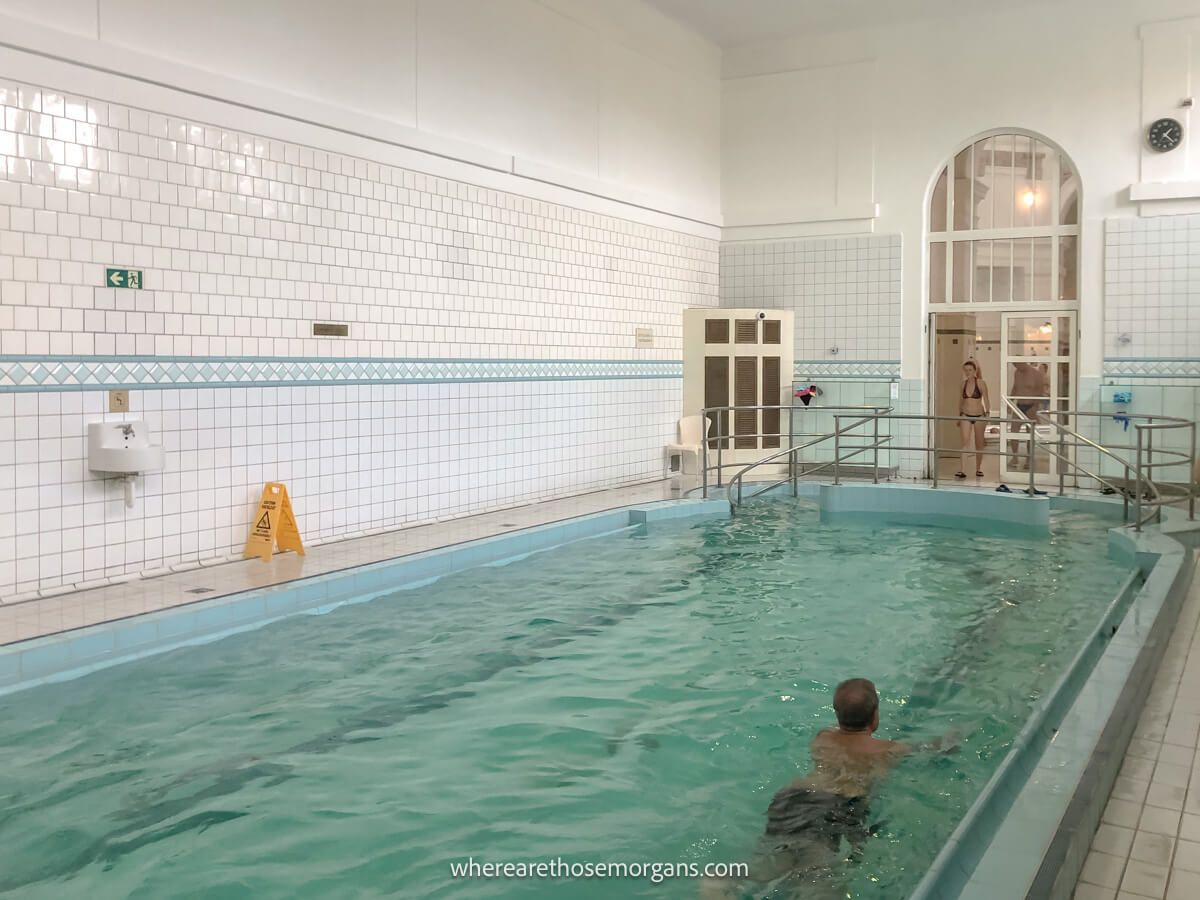 Bather slowly swimming in a long indoor medicinal pool surrounded by white tiles