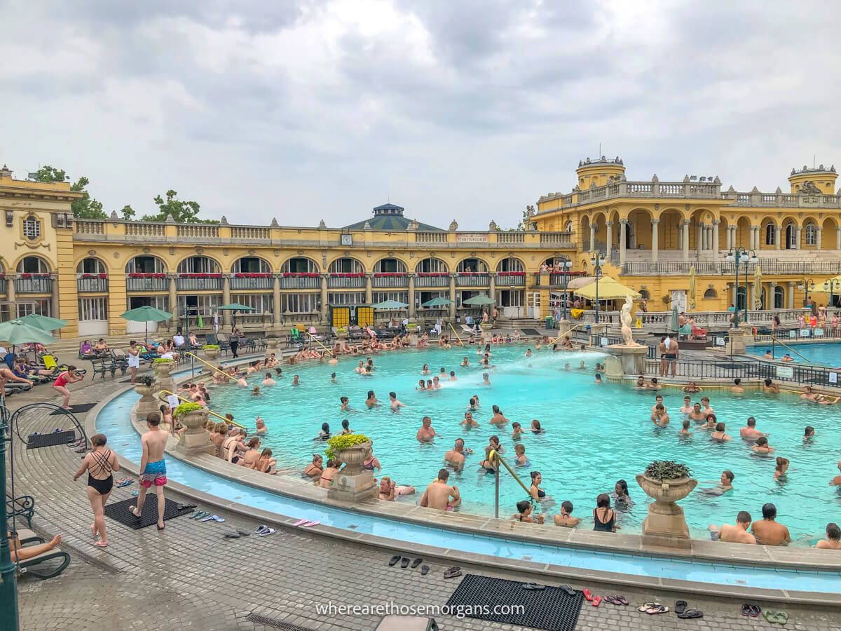 Photo of an outdoor thermal pool with lots of people bathing backed by a low and long yellow building on a cloudy day at Szechenyi Baths in Budapest