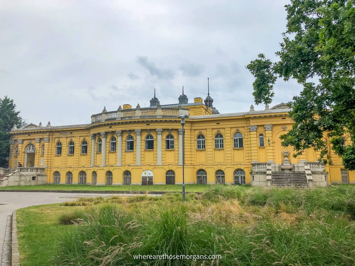 Photo of a large and long yellow building with old world architecture behind trees and grass on a cloudy day