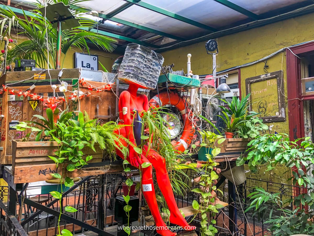 Photo of a red mannequin sat on a metal fence surrounded by plants inside a ruin bar in Budapest