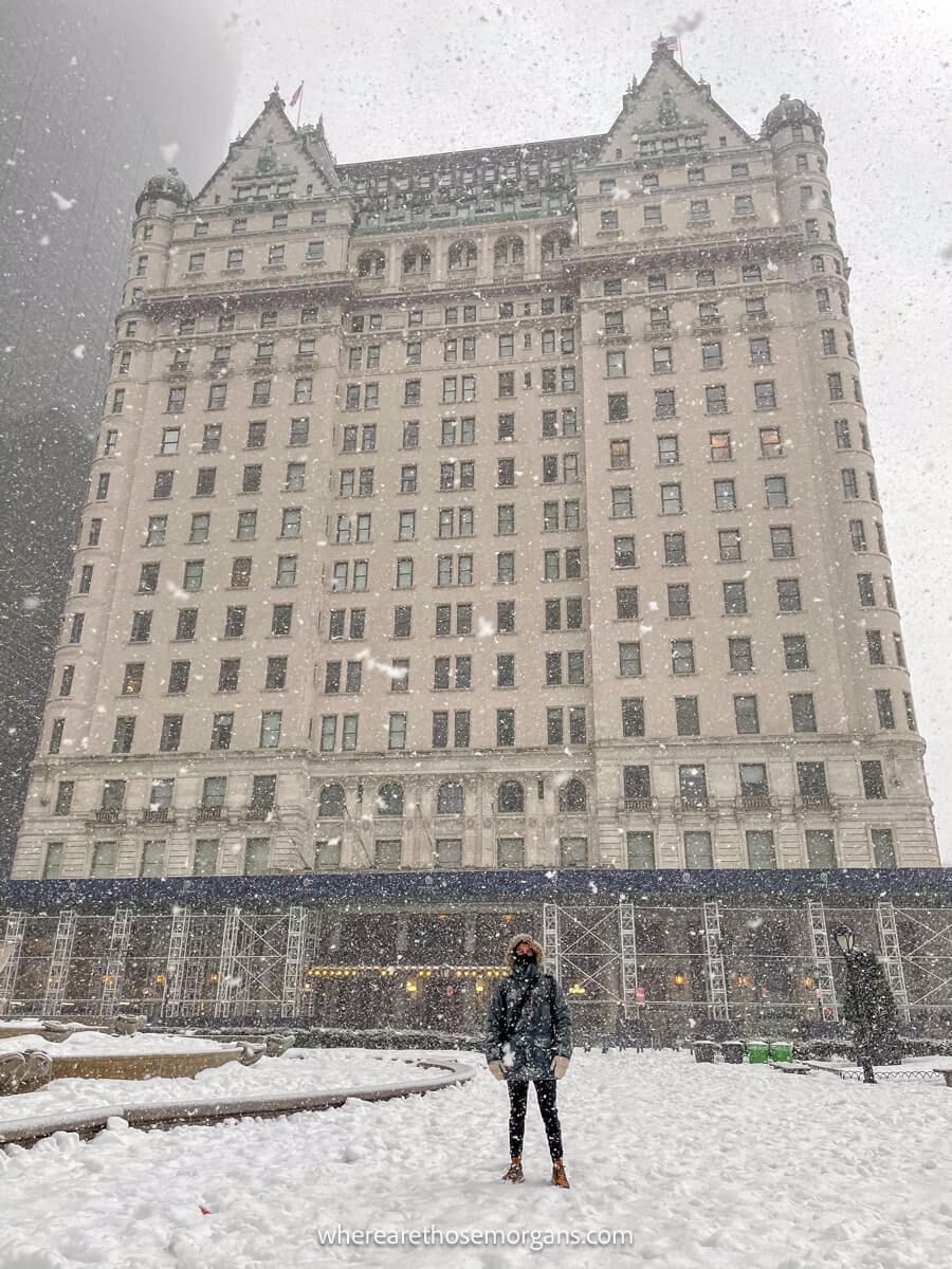 Photo of a tourist in thick coat standing outside a tall and wide hotel on a very snowy day
