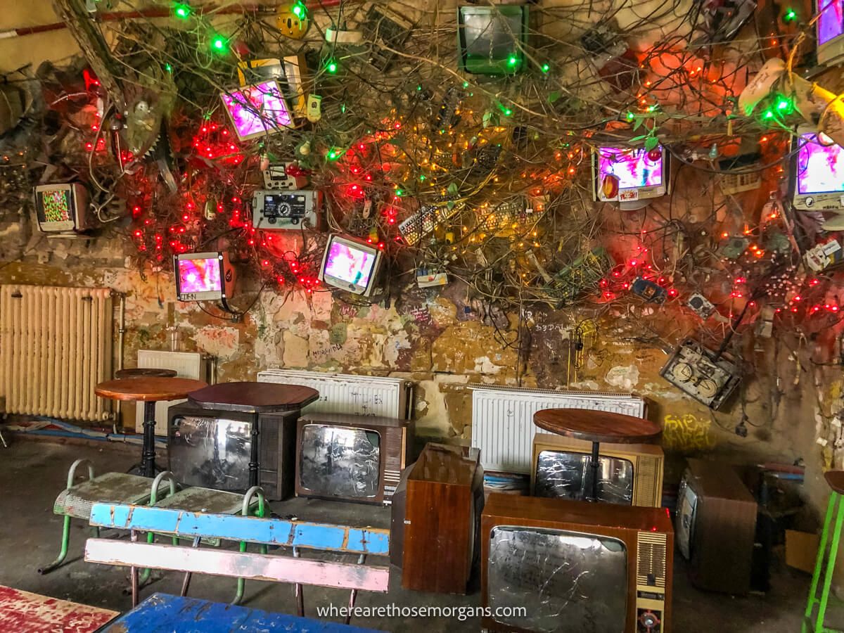 Lots of small computer monitors on a wall tangled with long cords and colorful fairy lights, and smashed up tv's on the ground in a ruin bar called Szimpla Kert in Budapest