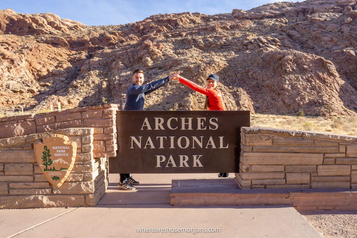 Photo of a couple standing behind an Arches National Park sign with arms joined together in the shape of an arch