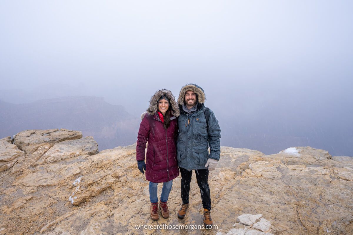 Photo of a couple standing together in heavy winter coats on a rocky ledge with long drops into a canyon behind but it's shrouded in snow clouds
