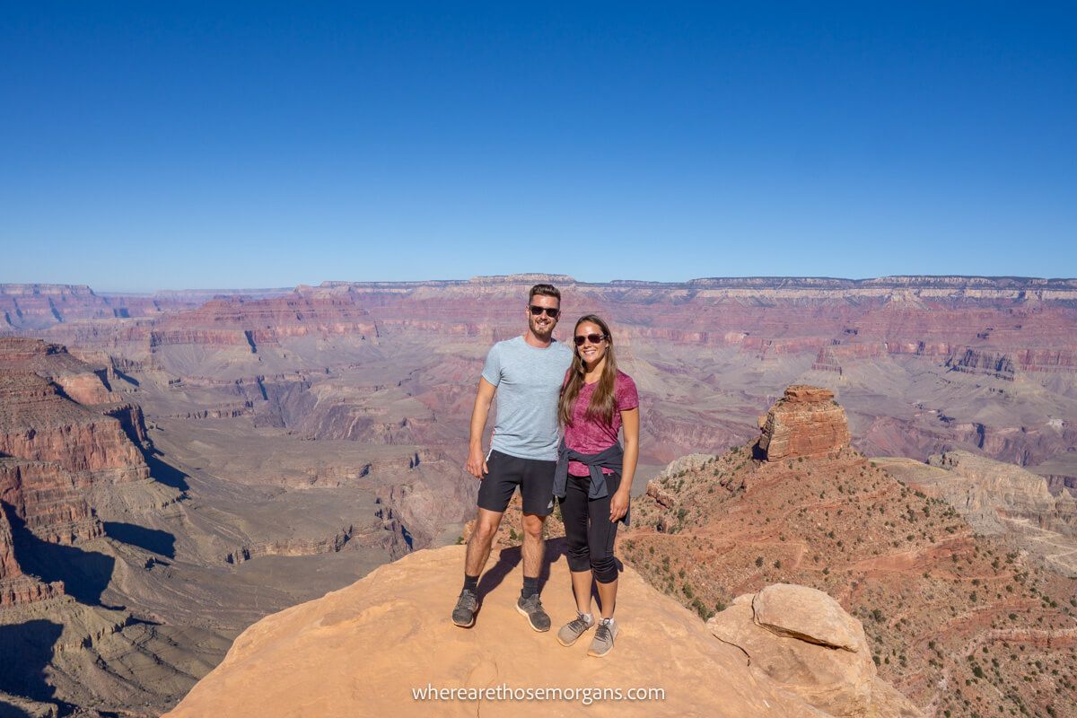 Photo of a couple standing together on a rocky ledge with far reaching views over a wide open rocky landscape on a sunny day