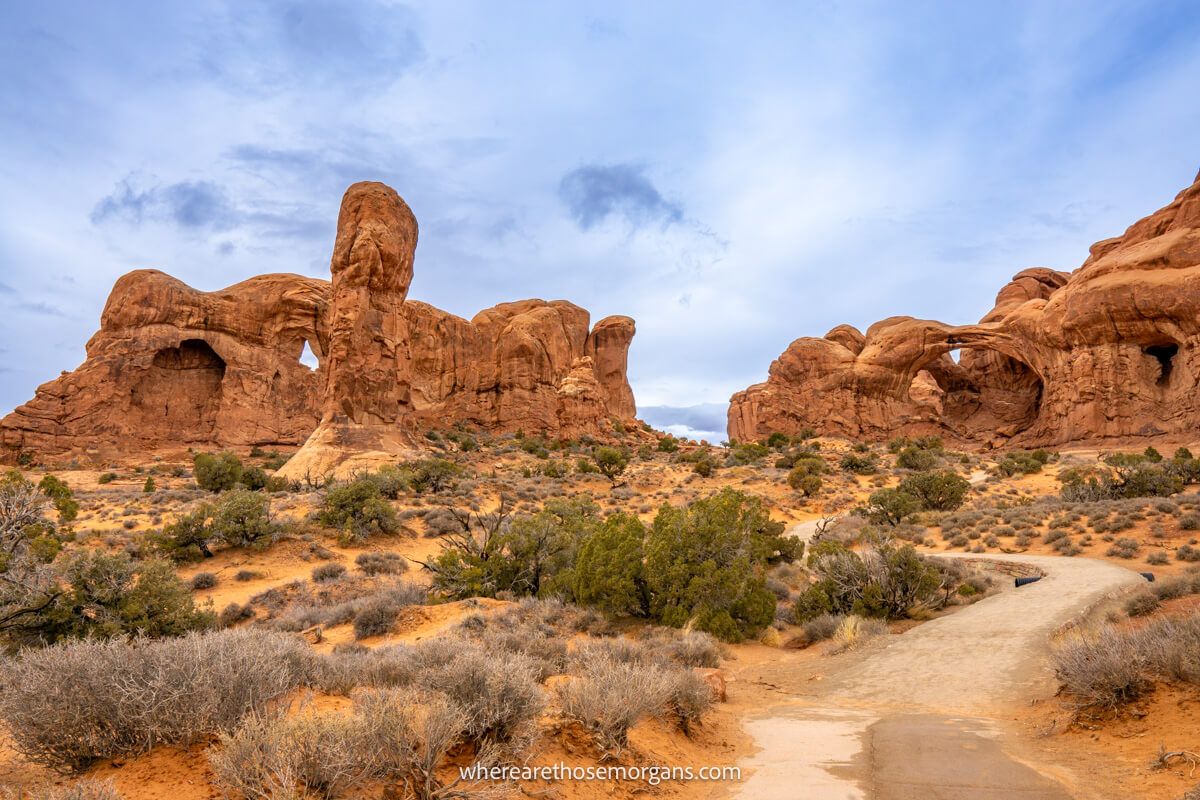 Photo of a path leading through desert vegetation to a cluster of oddly shaped red rock formations under a cloudy sky