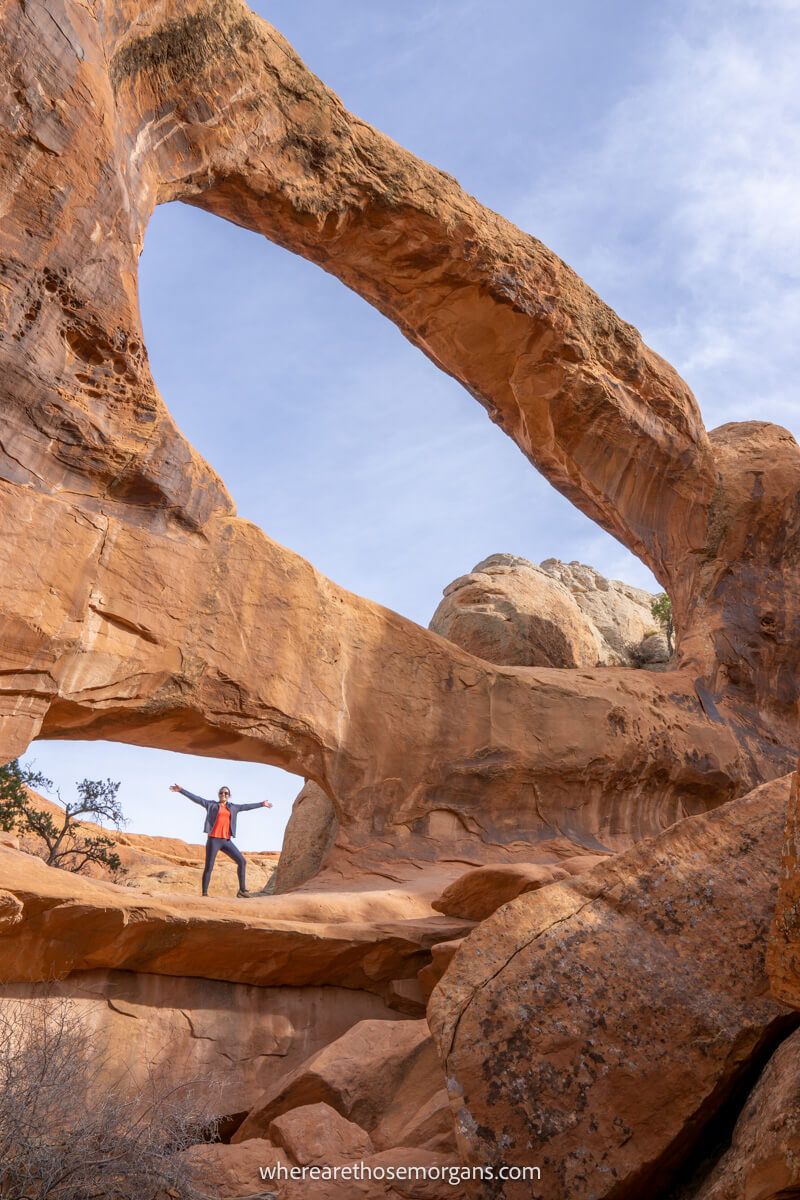 Hiker stood with arms in the air inside a window shaped hole in a red rock formation with another hole above on the Devils Garden Trail in Arches National Park