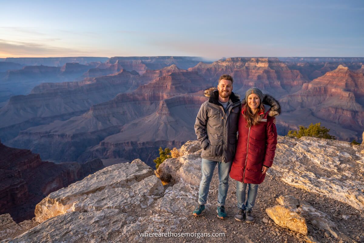 Photo of Mark and Kristen Morgan from Where Are Those Morgans standing together in winter coats on a rocky ledge with far reaching views over Grand Canyon South Rim filled with red rock formations at sunset