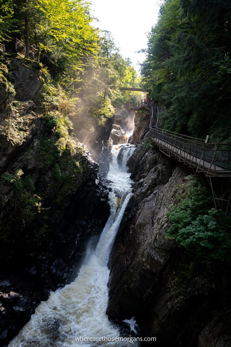 Photo of High Falls Gorge in Lake Placid with waterfalls crashing through a narrow gorge next to a wooden boardwalk and sunlight bursting in from above