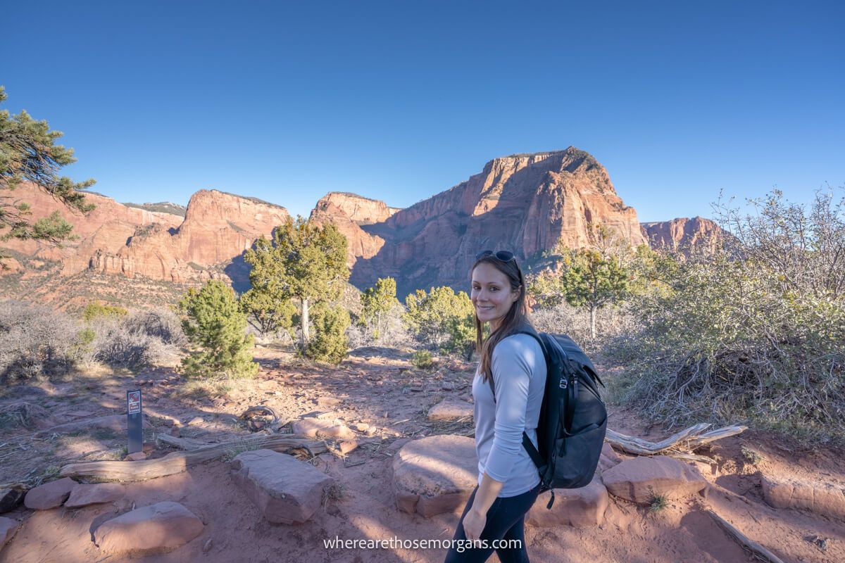 Photo of Kristen Morgan from Where Are Those Morgans hiking a dirt packed path with desert vegetation and red rock mountains on the Timber Creek Overlook Trail on a sunny day in Zion