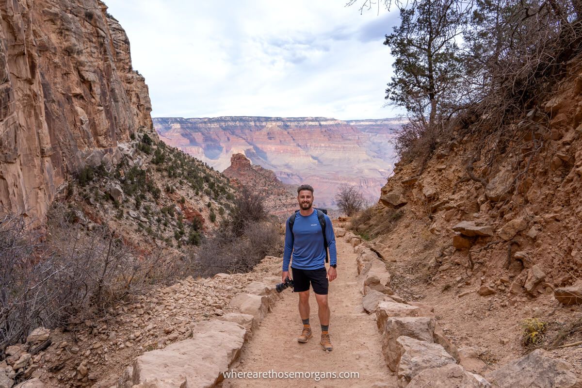 Photo of Mark Morgan hiking Bright Angel Trail in the Grand Canyon in shorts and a blue shirt standing on a narrow path with red rocks all around and far reaching views into the canyon behind