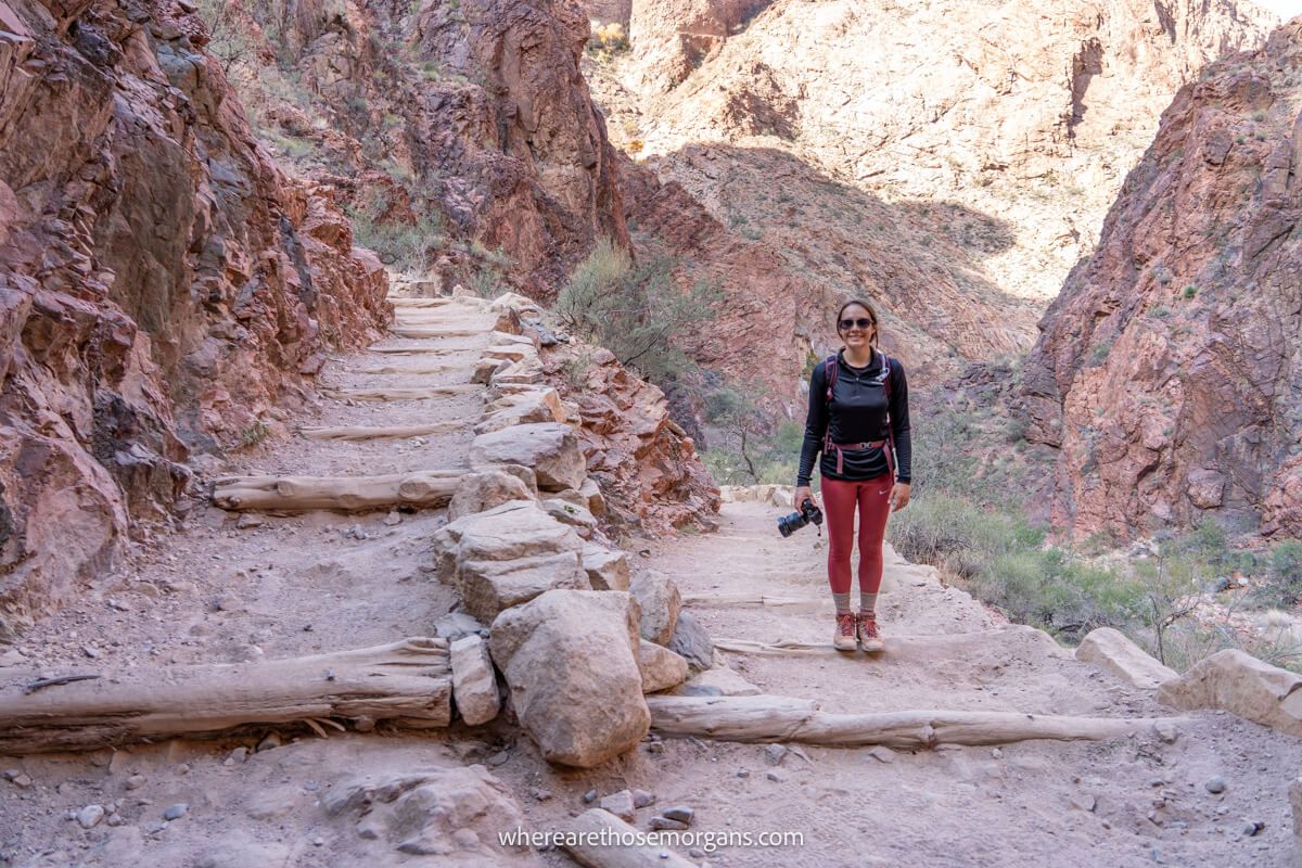 Photo of Kristen Morgan from Where Are Those Morgans in red pants and black shirt holding a camera standing on a tight switchback hiking Bright Angel Trail in the Grand Canyon