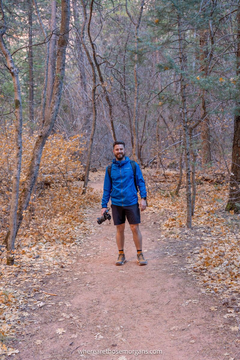 Photo of a hiker in a blue coat holding a camera in a light forest on a dirt trail
