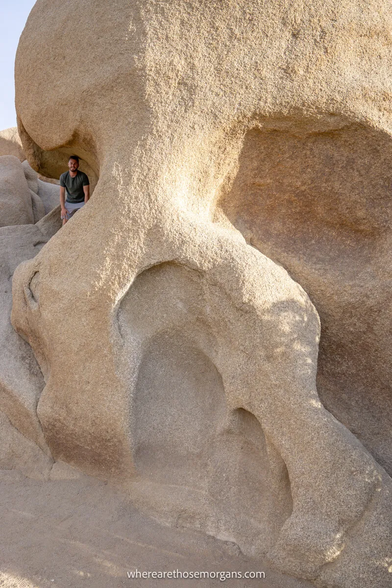 Hiker crouched in a small depression in a rock formation that looks a bit like a skull in Joshua Tree National Park
