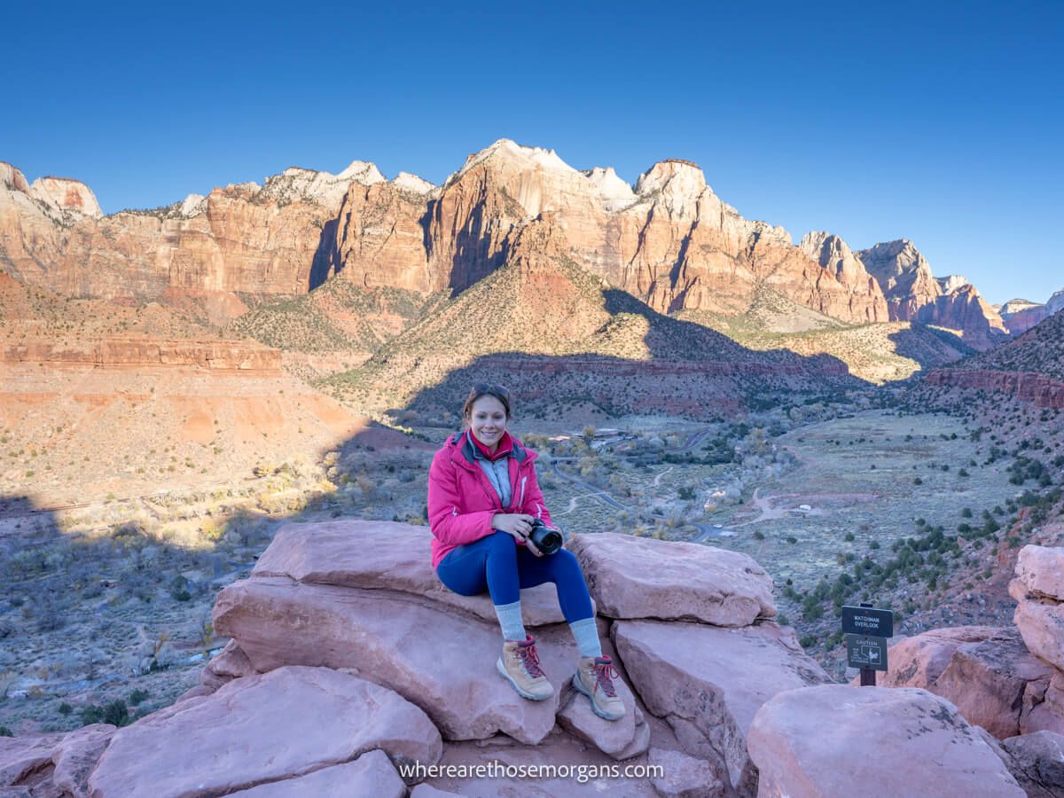 Photo of a hiker sat on rocks with winter coat at sunrise in Zion enjoying far reaching views over a wide open valley behind