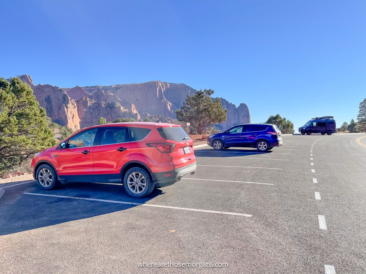Photo of a parking lot with 3 cars and red rock mountains behind on a sunny day
