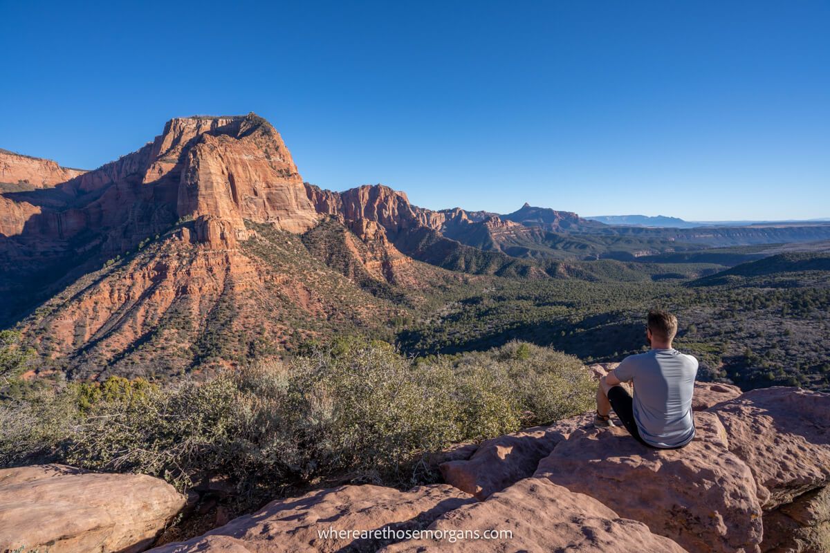 Photo of a hiker sat on red rocks looking at far reaching views over a deep valley filled with trees and tall red rock mountains under a clear blue sky at Timber Creek Overlook in Zion National Park