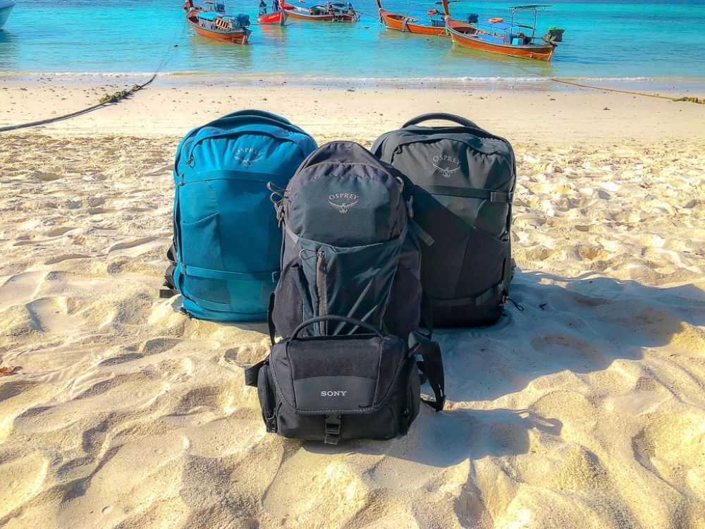 Three osprey backpacks and a camera bag sitting on a beach in Thailand