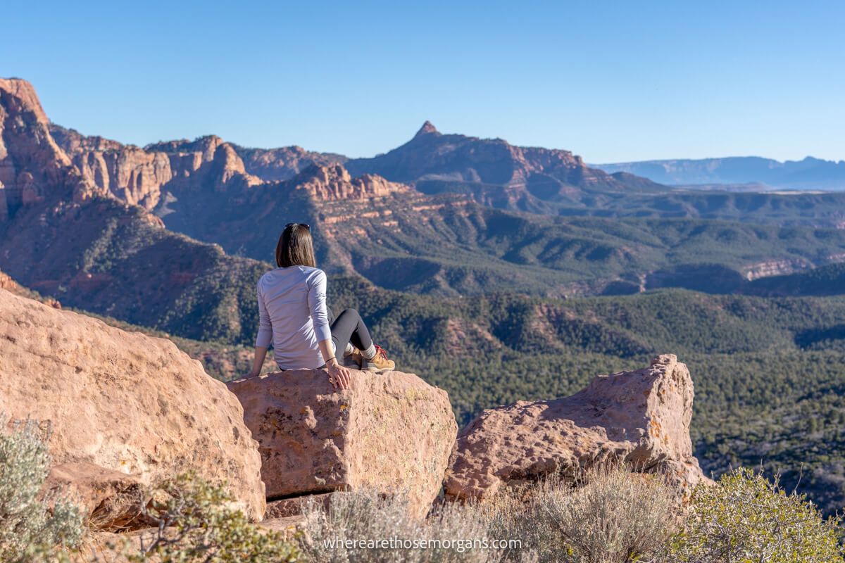 Photo of a hiker sat on large red boulders looking out at a wide open view of red rocks and trees in a deep valley at Timber Creek Overlook in Zion