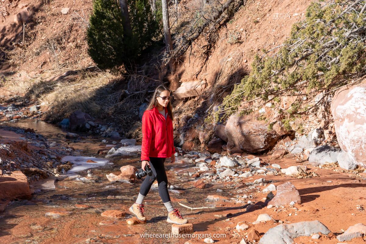 Photo of a hiker walking across stones over a shallow creek holding a camera with the sun warming up on a cold morning