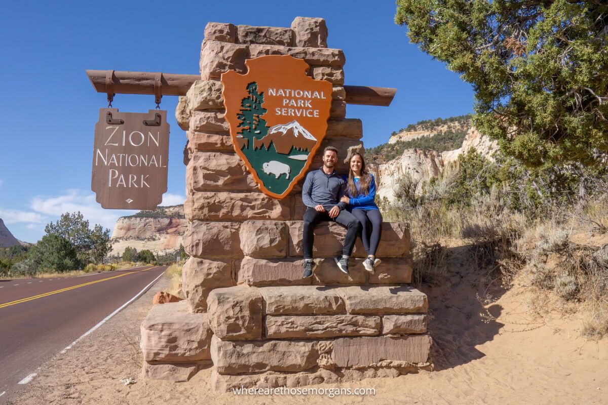 Photo of Mark and Kristen Morgan from Where Are Those Morgans sitting on a rock wall underneath the entrance sign for Zion National Park next to the road on a sunny day