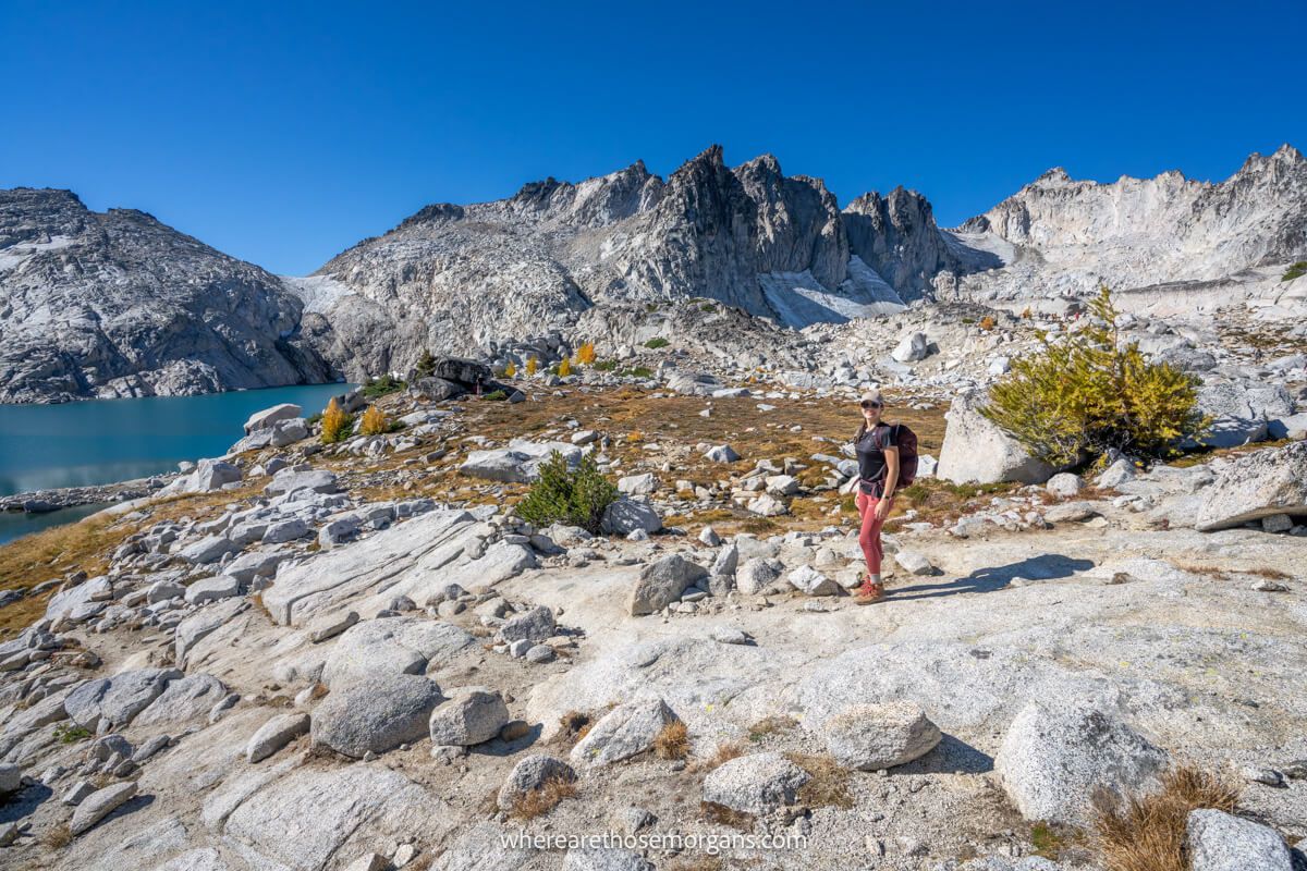 Photo of a hiker stood on a rocky trail with a lake and rugged granite mountains behind under a deep blue sky