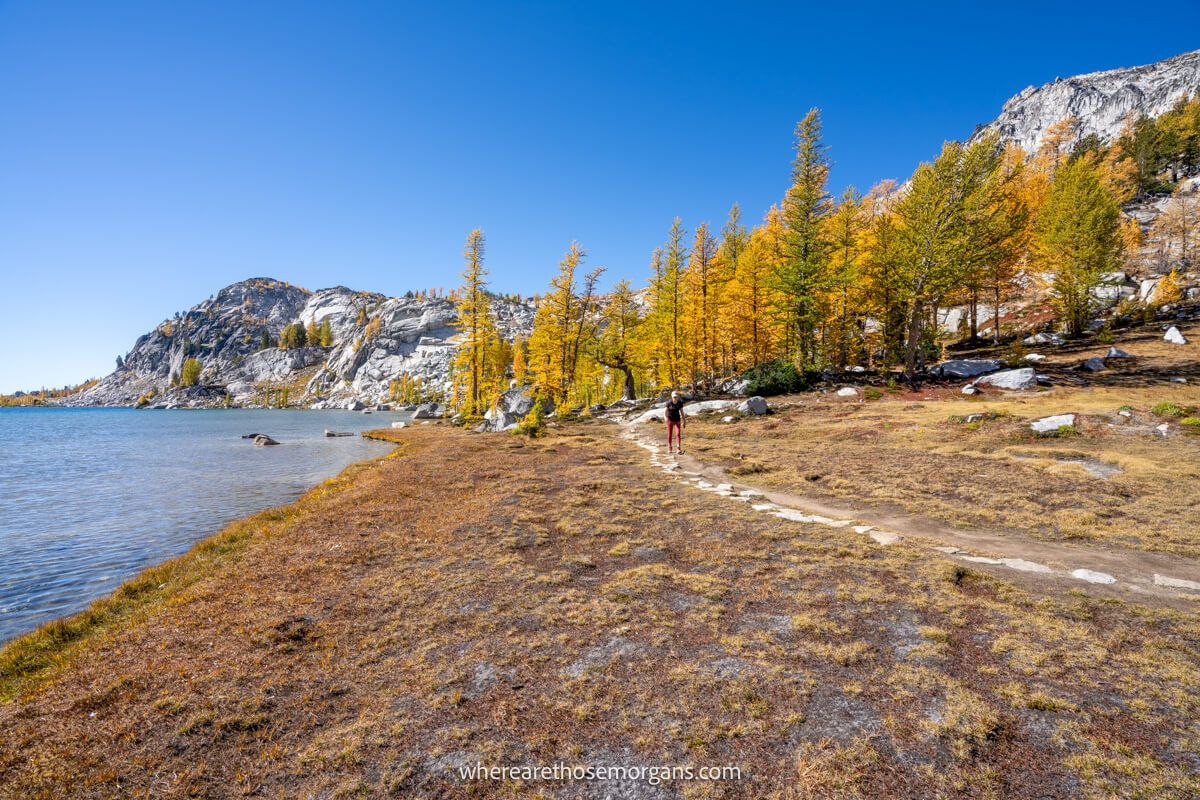 Photo of a person hiking along a narrow path next to a lake with golden larches and granite peaks behind on The Enchantments hike in Washington