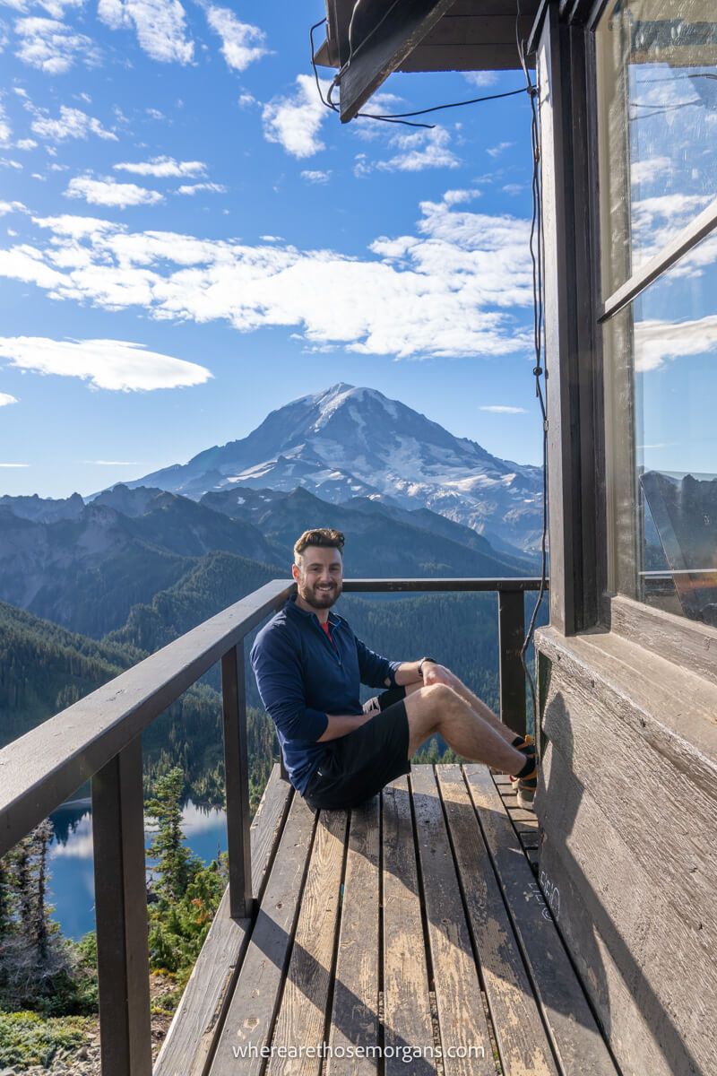 Photo of Mark Morgan from Where Are Those Morgans sat on the side of Tolmie Peak fire lookout tower with Mt Rainier behind on a sunny day