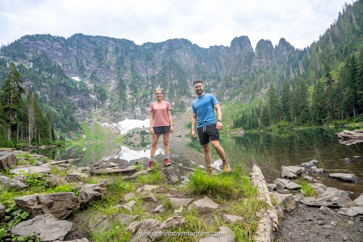 Photo of Mark and Kristen from Where Are Those Morgans standing in shorts and t-shirts on small rocks on the edges of Lake Twentytwo with tall mountain cliffs behind and green tree leaves all around