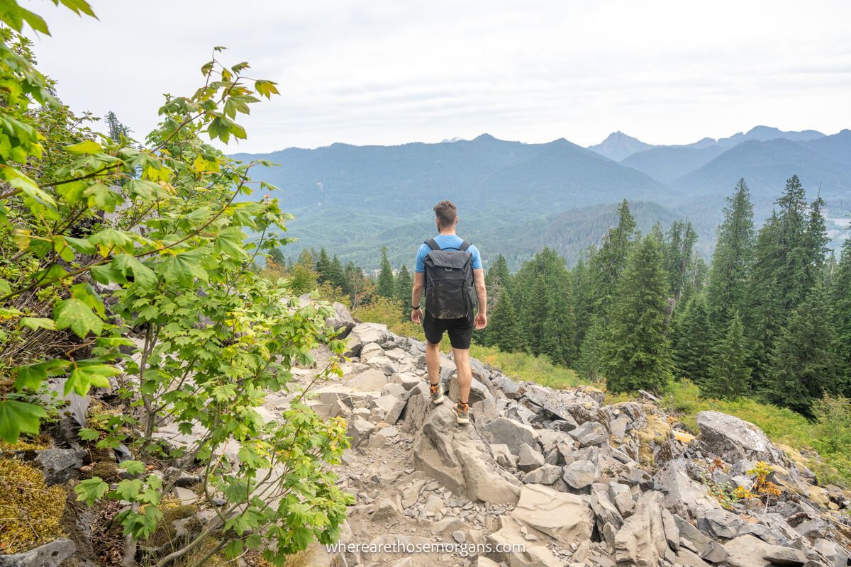 Photo of a person walking on small rocks on a mountainside looking at views over rolling hills and trees on a cloudy day in Washington State