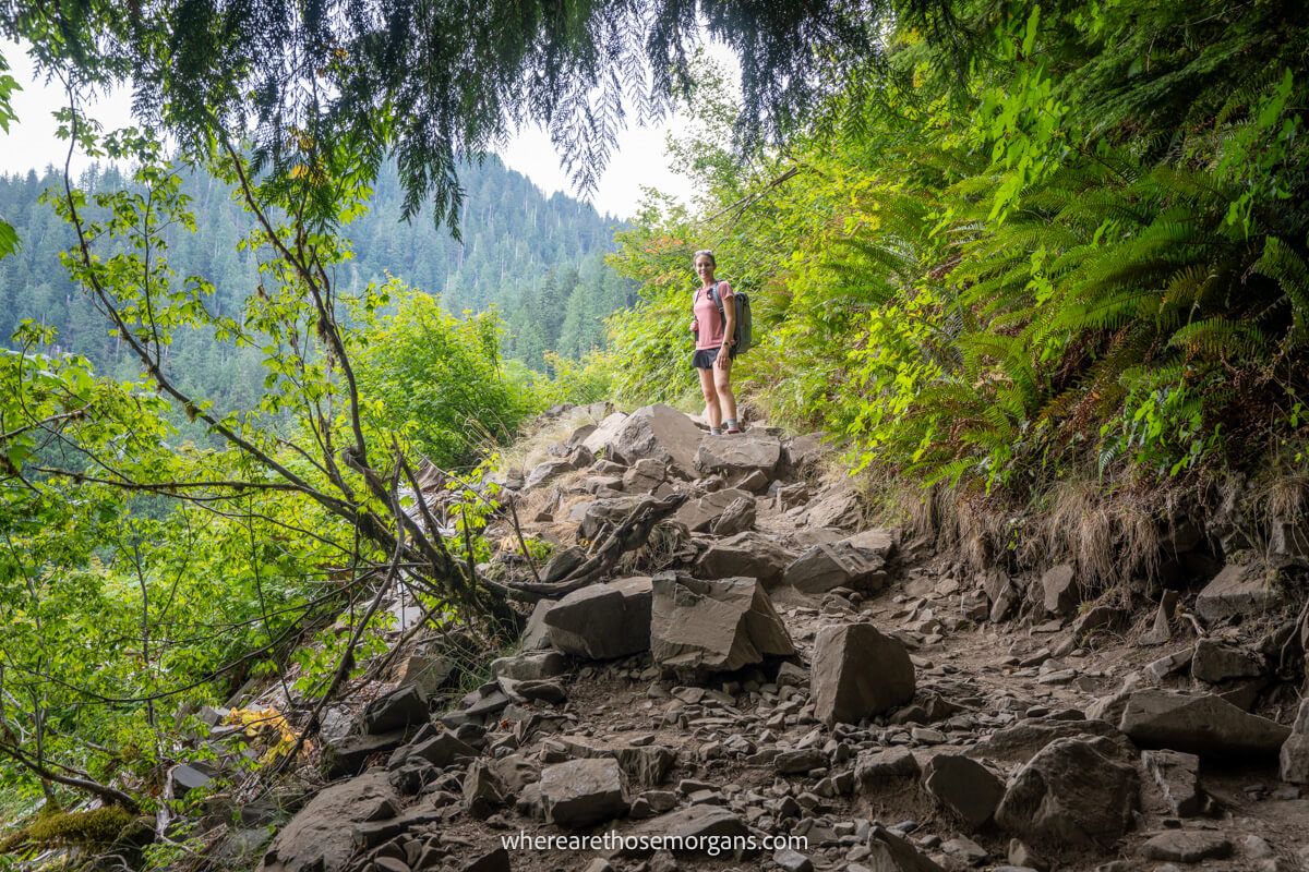 Photo of Kristen from Where Are Those Morgans in pink t-shirt with backpack stood on top of small rocks on a rocky hiking trail next to trees and vegetation on the way to Lake 22 in Washington