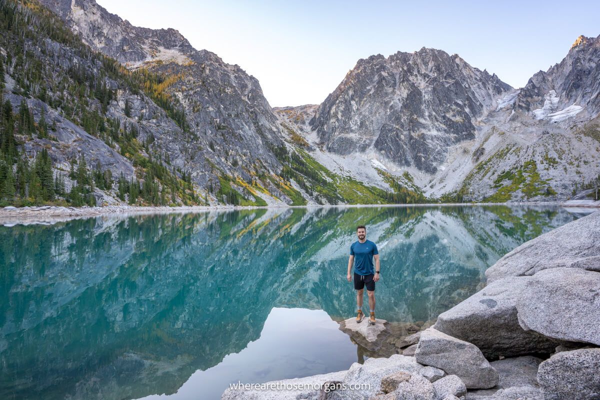 Photo of Mark Morgan stood in shorts and t-shirt on a rock on the shoreline of Colchuck Lake surrounded by tall rugged mountains on The Enchantments hike in Washington at dawn