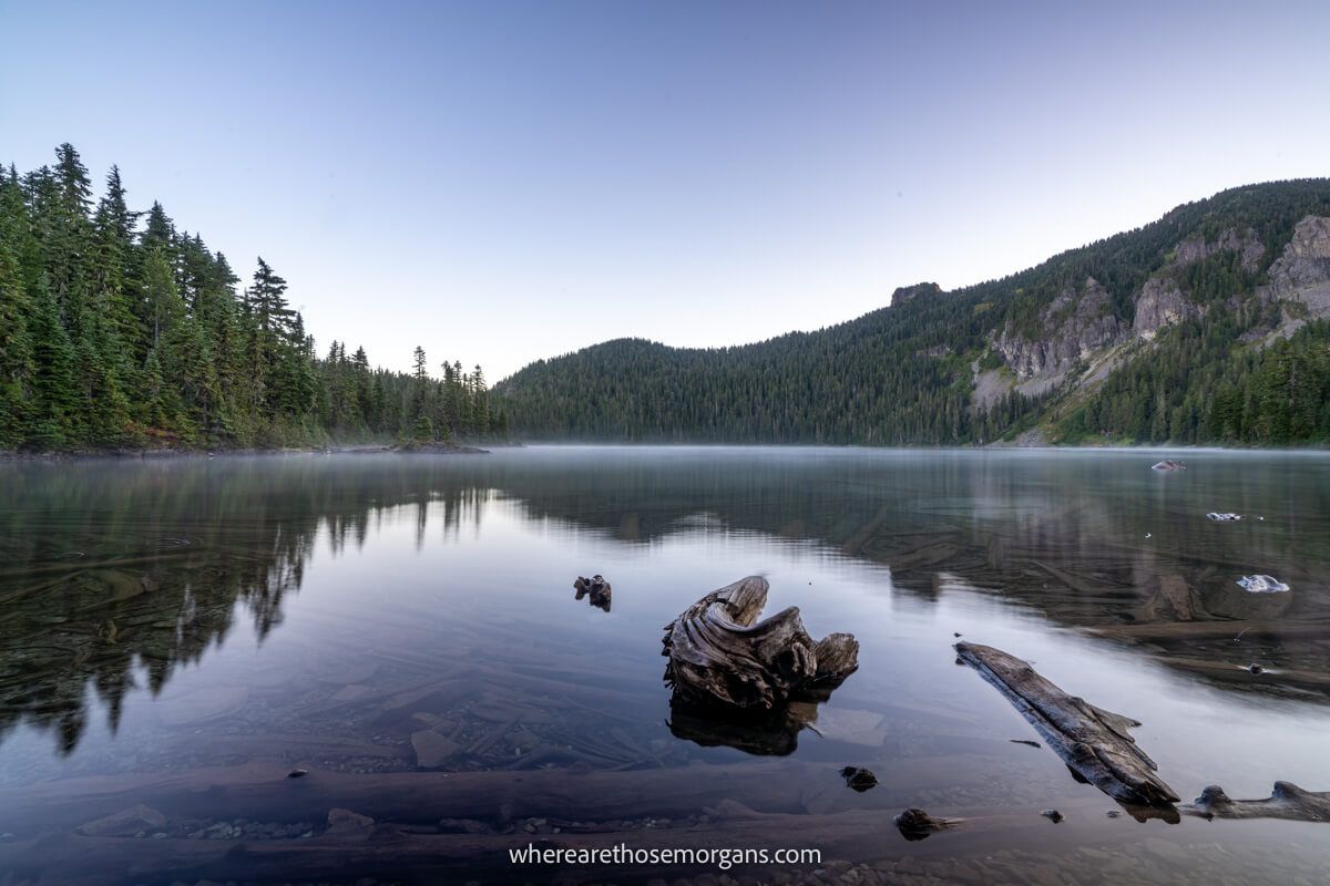 Photo of a large and perfectly still lake with a thin veil of mist on the flat surface, along with trees and a mountain side reflecting