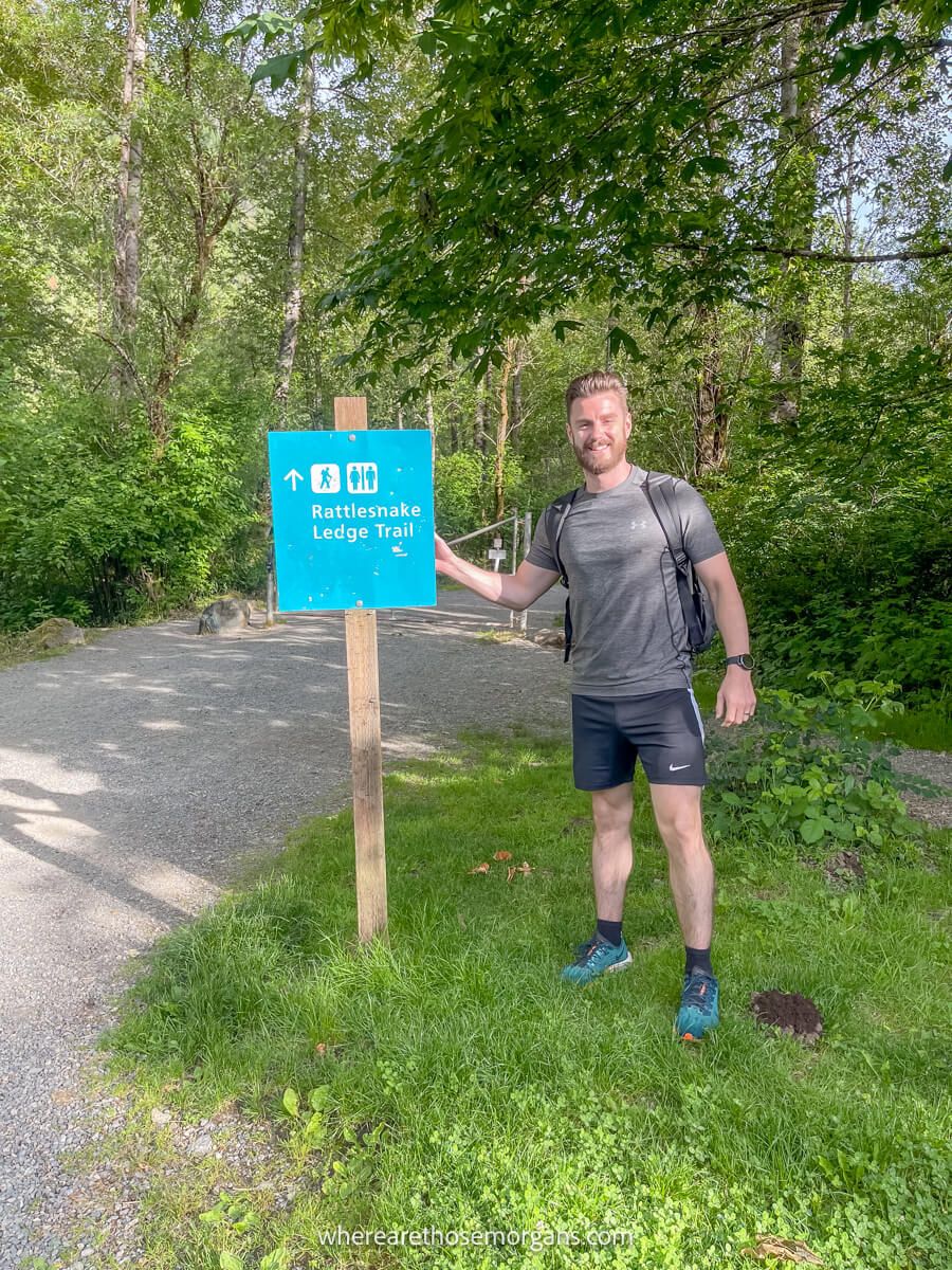 Photo of a hiker stood next to a wooden trailhead sign with trees behind