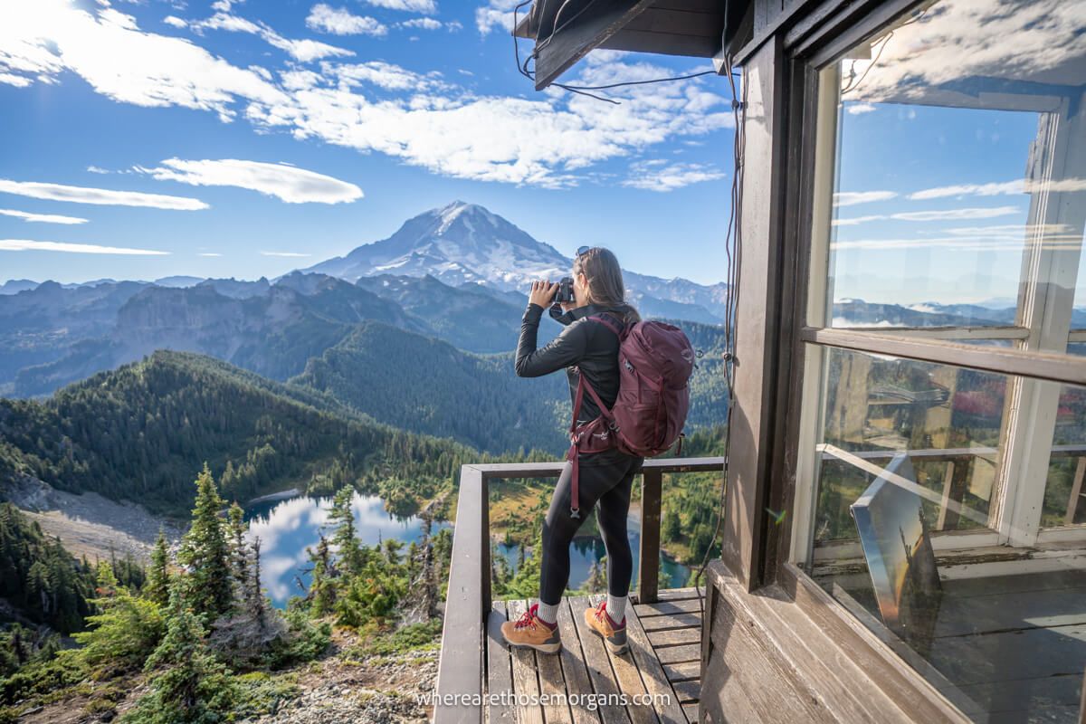 Photo of a hiker taking photos from a wooden fire lookout tower overlooking a lake and volcano at the top of Tolmie Peak