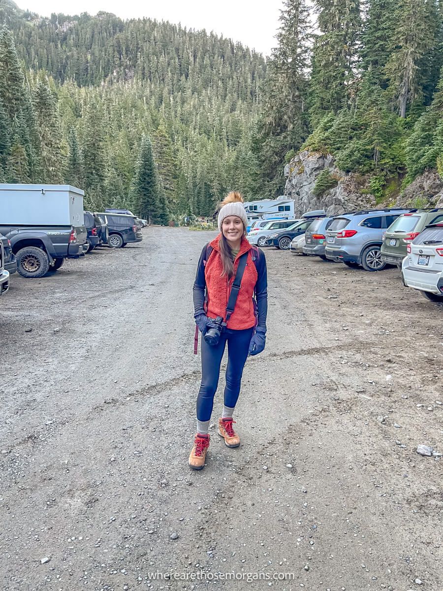 Photo of a hiker standing in winter gear in a gravel parking area with cars parked on either side of the road at Tolmie Peak Trailhead in Washington