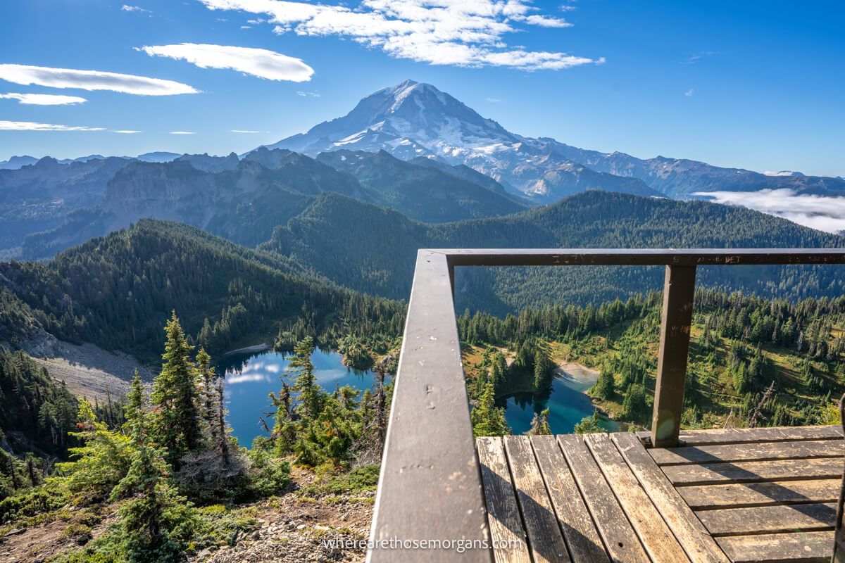 Photo of Mount Rainier taken from Tolmie Peak fire lookout tower with a wooden rail leading to a lake, trees and snow capped volcano on a clear sunny day
